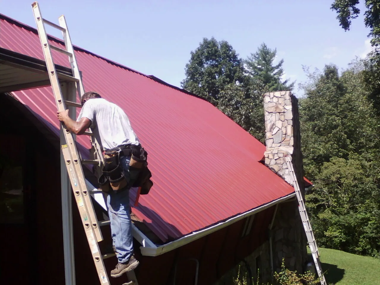 Roofer on a ladder beside a red metal roof and stone chimney — Soffit & Fascia Repair by Roofing Brothers in Wood River